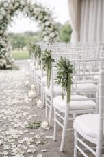 Allée pour célébration du mariage Chaises blanches décorées de verdure, allée de pétales et arceau floral en arrière-plan.